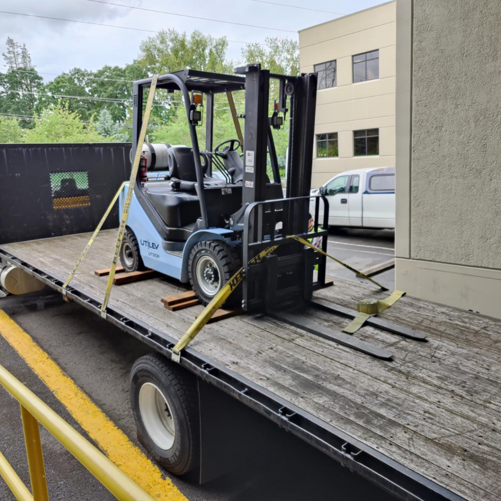 A blue forklift securely attached to the back of a truck and is getting dropped off at the development store. 