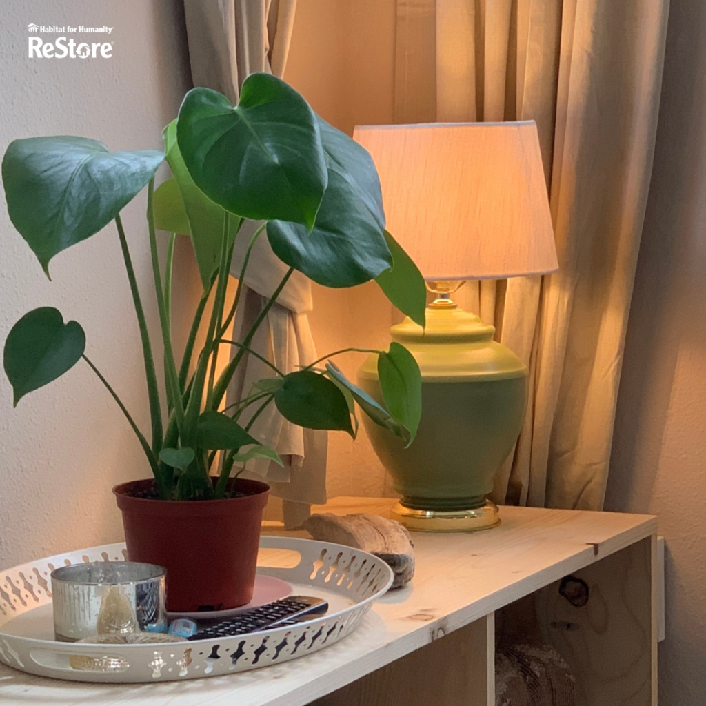 A turned on green lamp is on a shelf along with a plant that's on a tray with a candle, remote, and tied up curtains behind the lamp. 